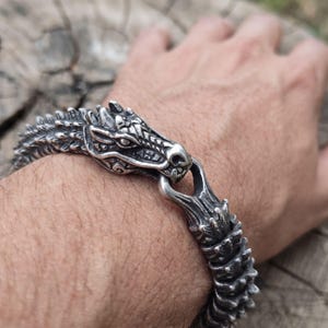 May include: A silver-toned metal bracelet featuring a detailed dragon head clasp. The band consists of articulated, scale-like segments that wrap around the wrist, with the dragon head connecting to the opposite end through a loop mechanism.