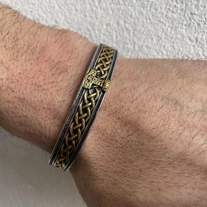 Può includere: Un bracciale argentato e dorato con un design di ispirazione vichinga. Il bracciale presenta un motivo intrecciato e un'emblema a martello color oro. Il bracciale è indossato al polso.