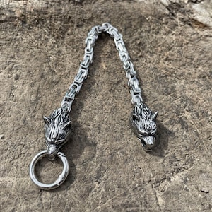 May include: A silver-colored chain accessory featuring two wolf head pendants. The pendants are connected by a chain link design. One wolf head has a circular ring attached. The accessory is displayed on a textured, brown surface.