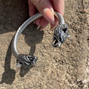 May include: Silver-toned cuff bracelet featuring wolf head details at each end. The bracelet has a twisted rope design and is held by a hand against a wood-grain background. The wolf heads have detailed fur and open mouths.