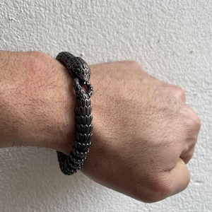 Op de afbeelding: Een donkergrijze metalen armband in de vorm van een slang, gewikkeld om een pols. De kop van de slang vormt de sluiting en het lichaam is gemaakt van overlappende schubben. De armband is een sieraad.