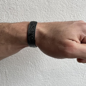 Könnte beinhalten: Ein schwarzes Armband mit einem aufwendigen Blumenmuster. Das Armband wird an einem Handgelenk getragen, wodurch sein detailliertes Design und seine dunkle Farbe hervorgehoben werden. Das Schmuckstück ist ein stilvolles Accessoire.