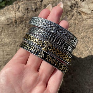 Può includere: Una mano mostra diversi braccialetti in stile vichingo argentati e dorati. I braccialetti presentano intricati motivi a nodi e iscrizioni runiche. I braccialetti sono impilati, mostrando i loro diversi disegni e colori.