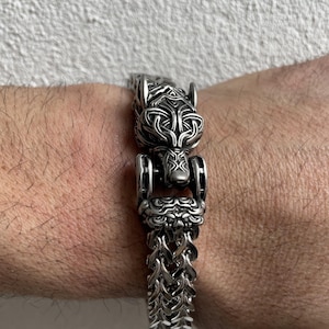 May include: A silver-colored bracelet featuring a detailed wolf head clasp. The bracelet has a chain-link design and is worn on a wrist. The wolf head clasp has intricate carvings and is the focal point of the jewelry.