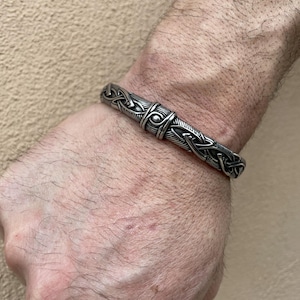 Puede incluir: Un brazalete plateado con intrincados diseños de nudos celtas y geométricos. El brazalete se lleva en la muñeca, mostrando el detallado trabajo en metal y la estética general de la joya.
