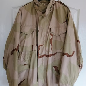 May include: A desert camouflage jacket in shades of tan and brown, featuring multiple pockets and a high collar. The jacket is suspended on a black hanger against a white door. It has a button-up front and a zip closure.