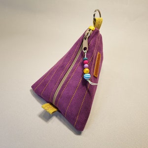 May include: A purple triangular zippered pouch with a yellow zipper pull and a beaded keychain with colorful beads. The pouch is made of a textured fabric.