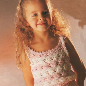 Pode incluir: Uma menina usando um top e uma saia de malha rosa e branca feitos à mão. O top apresenta um delicado padrão floral e um decote com vieiras. Uma flor branca adorna seu cabelo. A roupa é para uma criança.