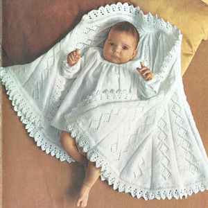 May include: A white crocheted baby blanket with a delicate lace trim. The blanket is folded around a baby wearing a white dress.