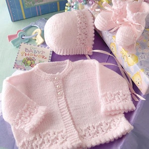 May include: A light pink knitted baby cardigan with three pearl-like buttons, a matching bonnet with ribbon ties, and a pair of knitted booties with ribbon bows. A small square card with the text "for BABY'S SHOWER" is visible nearby.