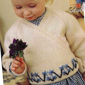 May include: A white knitted baby cardigan with a blue and white checkered pattern on the bottom. The cardigan has a V-neck and a blue embroidered design on the bottom.