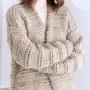 May include: A beige knitted cardigan with a long, open front and a relaxed fit. The cardigan is made of a chunky yarn and has a textured stitch pattern.