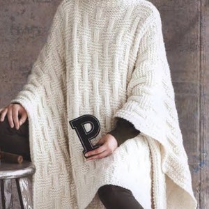 May include: A white knitted poncho with a turtleneck collar and a cable knit pattern. The poncho is worn by a person sitting on a stool.
