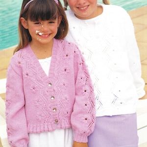 May include: Two young girls smiling, wearing knitted sweaters. One girl wears a light pink cardigan with flower details and button closures. The other girl wears a white sweater with a diamond pattern. Both sweaters are handmade.