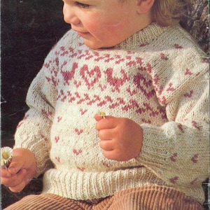 May include: A young child wearing a cream-colored knitted sweater with a pink and white geometric pattern. The sweater has a round neckline and long sleeves. The child is wearing brown corduroy pants.