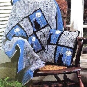 Puede incluir: Una manta y una almohada de ganchillo en azul y gris con la silueta de un gato en una ventana. La manta está colocada sobre una mecedora de madera.