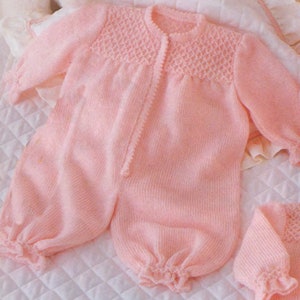 Puede incluir: Un pelele de punto rosa para bebé con cierre de botones y puños con volantes. El pelele está hecho con un delicado patrón de punto y tiene un gorro a juego.