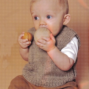 May include: A baby wearing a brown knitted vest, white shirt, and brown pants, holding and chewing on two wooden toys. The baby is sitting, with a neutral background.