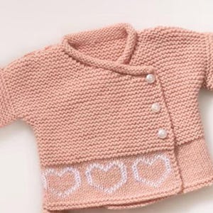 Puede incluir: Un cárdigan de bebé de punto rosa con detalles de corazón blanco y botones de perla.