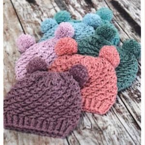 May include: A collection of six crocheted hats with pom-poms in various colors, including pink, purple, blue, and green. The hats are arranged in a row on a wooden surface.
