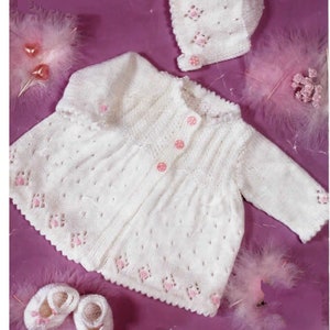 Puede incluir: Un cárdigan de punto blanco para bebé con botones rosas y un gorro a juego. El cárdigan tiene un delicado patrón de encaje y un borde festoneado. El conjunto es perfecto para un bebé recién nacido.