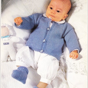 Könnte beinhalten: Ein Baby trägt einen hellblauen Strickcardigan mit Knöpfen, eine weiße Hose und blaue Strickschuhe. Das Baby liegt auf einer weißen Decke mit weißer Spitzenborte.