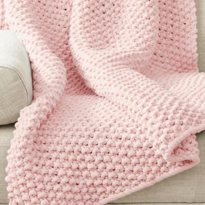 May include: A light pink knitted blanket with a textured, raised stitch pattern. The blanket is draped over a neutral-coloured surface, showcasing its cosy and inviting appearance. The thick knit suggests warmth and comfort.