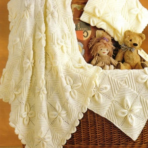 May include: A white knitted baby blanket with a floral pattern. The blanket is folded and displayed on a wicker basket with a teddy bear and a doll.