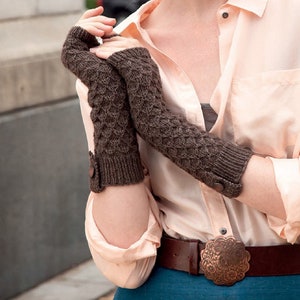 Peut inclure: Une paire de mitaines en tricot marron avec un détail bouton. Les mitaines sont portées par une personne portant une chemise boutonnée rose clair et une ceinture marron avec une grande boucle en métal.
