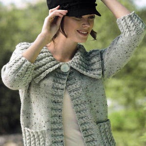 Puede incluir: Un cárdigan de punto gris claro con un cuello grande, cierre de botón y dos bolsillos. El cárdigan tiene un patrón moteado y se lleva con una camiseta blanca debajo.