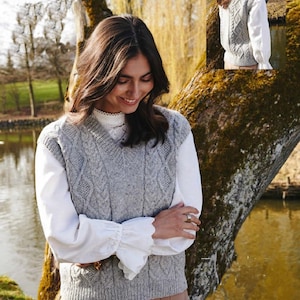 May include: A gray cable knit vest with a V-neck. The vest is worn over a white blouse with ruffled sleeves. The person is standing in front of a tree and a body of water.