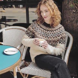 May include: A woman wearing a cream-coloured sweater with a geometric pattern in brown and white, seated at a cafe table. She holds a white cup, and the table is turquoise. The chair is white wicker.