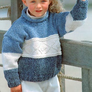 May include: A child's knitted jumper in shades of blue and white. The jumper features a textured diamond pattern across the chest and a ribbed turtleneck. The sleeves and lower section are a darker blue, creating a colour-block design.