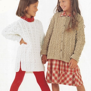 May include: Two young girls wearing knitted sweaters. The girl on the left is wearing a white sweater with a red scarf and red leggings. The girl on the right is wearing a brown sweater with a red and white plaid skirt.
