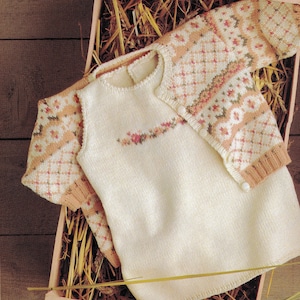 May include: A cream-colored knitted baby outfit, including a sleeveless top and a cardigan with a diamond and floral pattern in peach and brown. The outfit is displayed in a wooden box with straw.