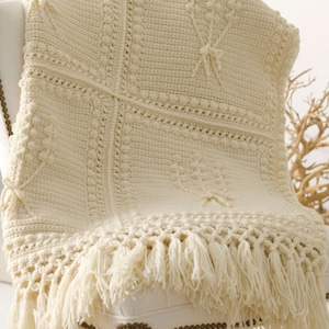 May include: A crocheted throw blanket in a cream color with a fringe border. The blanket is made with a textured stitch and has a delicate, feminine design.
