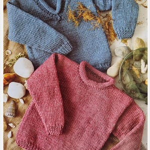 Peut inclure: Deux pulls tricotés. L'un est bleu poussiéreux avec un col, l'autre est rose-rougeâtre avec un col rond. Les deux pulls ont des manches longues et des poignets côtelés. Les pulls sont présentés sur une surface claire avec des coquillages et des éléments décoratifs.