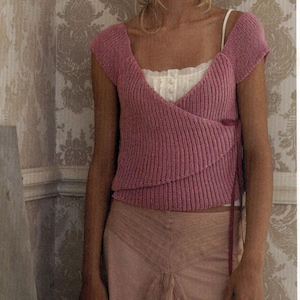 May include: A pink knitted wrap top with a tie at the side. The top is worn over a white camisole and a light pink skirt.