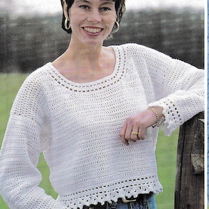 May include: A white crochet sweater with long sleeves and a scalloped hem. The sweater has a round neckline and is paired with blue jeans. The image shows a woman smiling and leaning on a wooden fence post.