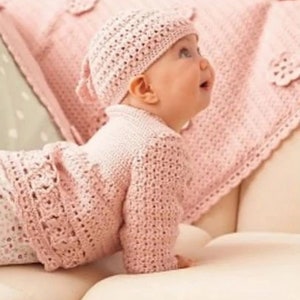 May include: A pink crocheted baby hat and cardigan set. The hat has a small bow on the back. The baby is wearing the set and is lying on a white couch.