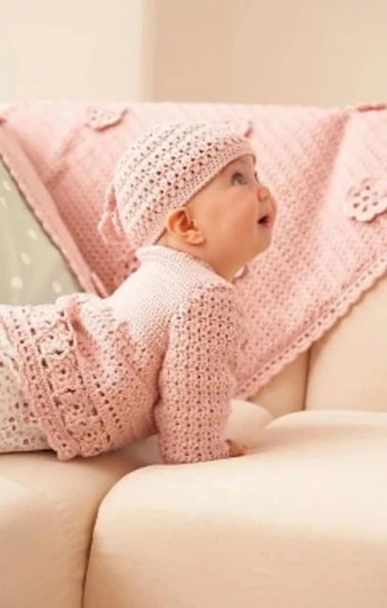 May include: A pink crocheted baby hat and cardigan set. The hat has a small bow on the back. The baby is wearing the set and is lying on a white couch.