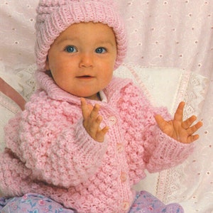 May include: A pink knitted baby cardigan with a button fastening and a matching pink knitted hat with a pom-pom on top.
