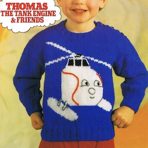 Peut inclure: Un pull en tricot bleu avec un motif d'hélicoptère blanc. L'hélicoptère a une bande rouge et un visage souriant. Le pull a le texte "THOMAS THE TANK ENGINE & FRIENDS" en haut.