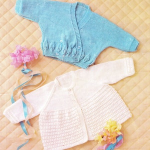 May include: Two knitted baby cardigans, one white and one light blue, laid out on a textured beige surface. The white cardigan has a button closure and a ribbed texture. The light blue cardigan has a wrap style with a leaf pattern and button closures.