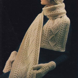 Könnte beinhalten: Ein weißes Strickset aus Mütze, Schal und Fäustlingen. Die Mütze hat einen Bommel oben. Der Schal ist lang und hat ein Zopfmuster. Die Fäustlinge sind fingerlos und haben ein Zopfmuster.