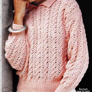 Puede incluir: Un suéter de punto rosa claro con cuello y mangas largas. El suéter presenta un patrón texturizado de tejido abierto. Las mangas y el cuerpo tienen un dobladillo acanalado. Se ve una pulsera plateada en la muñeca.