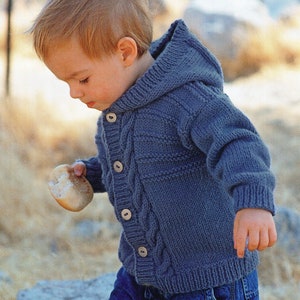 Może przedstawiać: Niebieski dzianinowy kardigan z kapturem i zapięciem na guziki. Kardigan ma wzór warkocza i jest noszony przez małe dziecko.