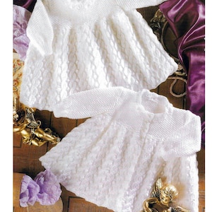 Puede incluir: Dos vestidos blancos de punto para bebé con un delicado patrón de punto de cable. El vestido de arriba tiene cuello alto y mangas largas, mientras que el vestido de abajo tiene un frente abotonado y un escote ligeramente más bajo.