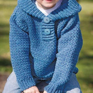 May include: A blue crochet hooded sweater with two large buttons. The sweater has long sleeves and a textured stitch pattern. The child is wearing blue jeans and a white shirt underneath the sweater.