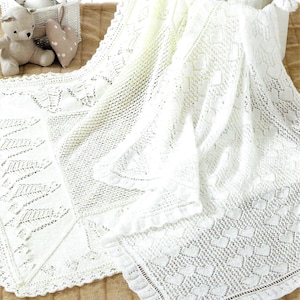 Peut inclure: Une couverture de bébé blanche, finement tricotée, avec un motif de dentelle délicat. La couverture présente une variété de motifs, notamment des cœurs et des formes géométriques. Les bords sont finis avec une bordure festonnée. Un ours en peluche et un oreiller en forme de cœur sont en arrière-plan.